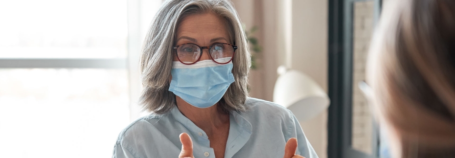A woman with gray shoulder-length hair speaks to another woman in an office. Both are wearing masks.